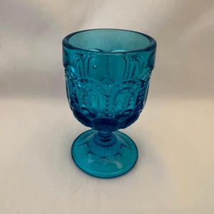 May include: A teal blue glass goblet with a decorative pattern. The goblet has a wide base and a round bowl.