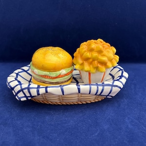 May include: A ceramic salt and pepper shaker set shaped like a hamburger and french fries. The hamburger has a yellow bun, a red tomato, and green lettuce. The fries are in a white container with orange stripes, all on a basket.
