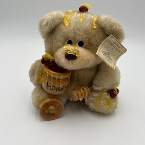 May include: A plush teddy bear with a honey-themed design. The bear is light brown with black eyes and a brown nose. It holds a honey pot with the word "HONEY" on it. The bear has honey dripping from its head and paws.