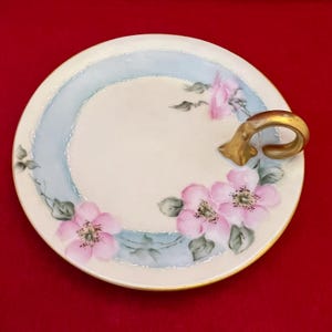 May include: A round, off-white serving plate with a gold handle. The plate features a light blue ring and is decorated with pink floral designs and green leaves. The rim has a gold trim.