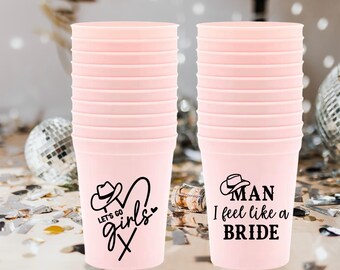 Può includere: Due pile di bicchieri di plastica rosa con testo e grafica neri. Una tazza dice "Let's Go Girls" con una grafica di cappello da cowboy. L'altra dice "Man I feel like a BRIDE" con una grafica di cappello da cowboy. Sfera disco e coriandoli sullo sfondo.