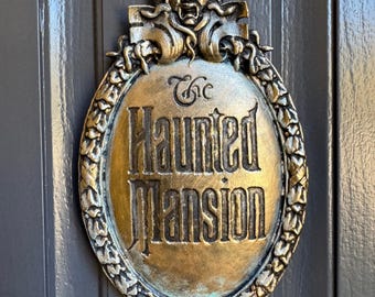 Könnte beinhalten: Ein ovales, bronzefarbenes Schild mit der Aufschrift "The Haunted Mansion" in kunstvollen Buchstaben. Das Schild ist von einem dekorativen Rahmen mit einem Totenkopf unten und einem grotesken Gesicht oben umgeben. Es ist an einer grauen Tür befestigt.