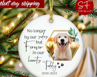 May include: A round white ceramic Christmas ornament featuring a photo of a golden retriever. The ornament has text that reads "No longer by our sides but forever in our hearts Teddy 2020-2023" with paw prints and floral accents.