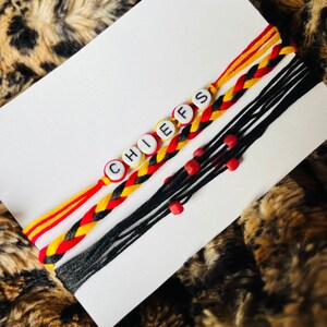Könnte beinhalten: Drei geflochtene Freundschaftsarmbänder mit rotem, gelbem und schwarzem Garn. Die Armbänder haben weiße Perlen mit den Buchstaben "CHIEFS" darauf.