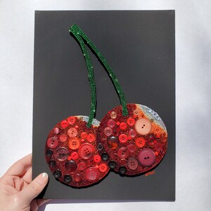 May include: A pair of red cherries made from buttons, with green beaded stems, on a black background.