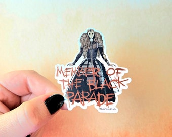 Könnte beinhalten: Ein Aufkleber mit einer Frau in einem schwarz-weißen Kleid mit einer Maske, die einen Blumenstrauß hält. Der Text "Member of the Black Parade" ist in Rot und Weiß auf dem Aufkleber geschrieben. Der Aufkleber wird in einer Hand mit schwarzem Nagellack gehalten.