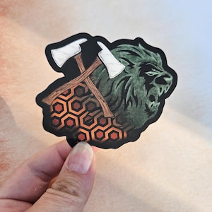 Peut inclure: Un autocollant noir et vert avec un graphique d'une tête de lion avec une paire de haches croisées derrière. La tête de lion grogne et a une expression féroce. Les haches sont en bois avec des lames blanches. L'arrière-plan est un motif d'hexagones orange et rouges.