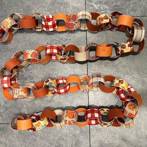 May include: A decorative paper chain featuring interlocking loops in various autumn-themed patterns and colors. The chain includes segments with orange, red and white buffalo plaid, pumpkin prints, and fall leaf motifs in shades of brown and gold.