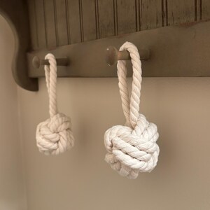 May include: Two off-white rope ornaments featuring a monkey fist knot design with a looped handle. Each ornament is constructed from thick, twisted cotton cord and hangs from a wooden peg, showcasing a textured, spherical knot at the base.