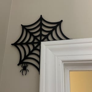 Può includere: Decorazione da parete in metallo nero a forma di ragnatela con un ragno appeso in basso. La ragnatela è fissata a uno stipite di porta bianco.