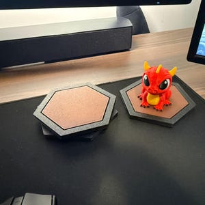 May include: A stack of two hexagonal coasters with black frames and textured brown centers, alongside a single matching hexagonal coaster featuring a small, red and yellow dragon figurine with large black eyes sitting on its brown surface.