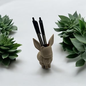 Puede incluir: Un organizador de escritorio de cerámica beige con forma de conejo con dos bolígrafos negros en su interior. El conejo tiene una textura esponjosa y está sentado sobre una superficie blanca.