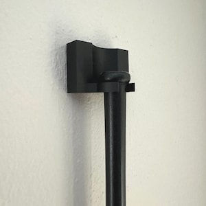 May include: A black, wall-mounted holder for a long, cylindrical object. The holder has a geometric design with a curved top and a small, round knob. The object is held in place by a small ledge.