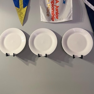 May include: Three white ceramic plates with a raised rim are mounted on a wall with black metal plate holders.