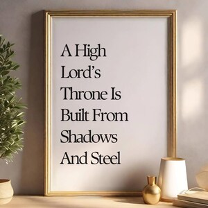 May include: A framed print with the text "A High Lord's Throne Is Built From Shadows And Steel" in black lettering. The frame is gold, and the print is displayed on a wooden surface with decorative items.