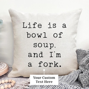 May include: A beige throw pillow with the text "Life is a bowl of soup, and I'm a fork" in black. The pillow has a neutral color and is placed on a surface with a decorative blanket and a vase with greenery.