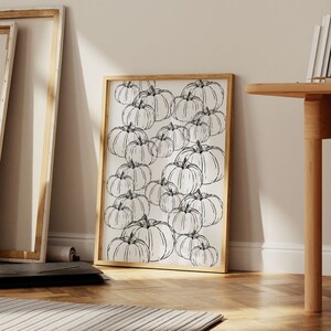 以下が含まれることがあります： 白黒の線画で描かれたカボチャが繰り返しパターンで描かれている。絵は明るい木製のフレームに額装され、白い壁に掛けられている。