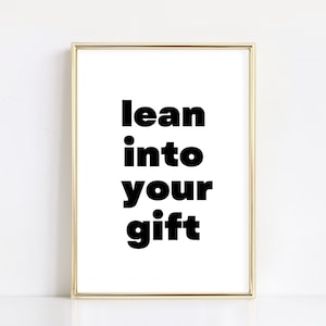 May include: A gold framed print with the text "lean into your gift" in black bold letters on a white background.