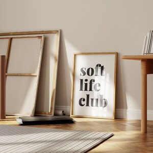 May include: A framed print with the words "soft life club" in black lettering. The print is in a light wood frame and leans against a wall. Other items in the room include a canvas, a rolled-up canvas, and a wooden table.
