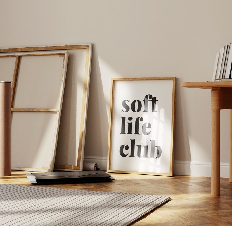 May include: A framed print with the words "soft life club" in black lettering. The print is in a light wood frame and leans against a wall. Other items in the room include a canvas, a rolled-up canvas, and a wooden table.