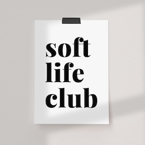 May include: A white poster with the words "soft life club" in bold, black font. The poster is held in place by a black clip at the top and is mounted on a white wall.