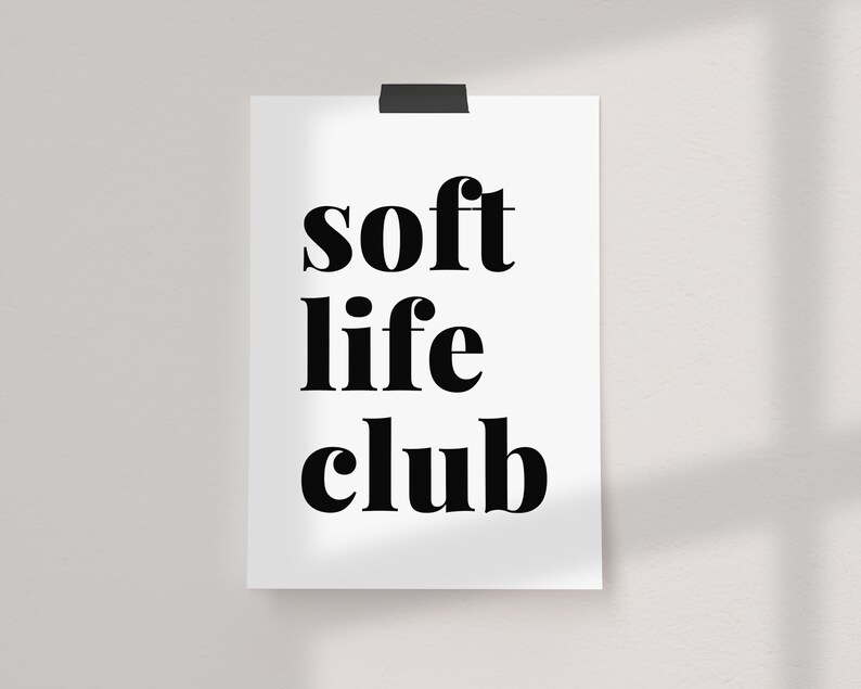 May include: A white poster with the words "soft life club" in bold, black font. The poster is held in place by a black clip at the top and is mounted on a white wall.