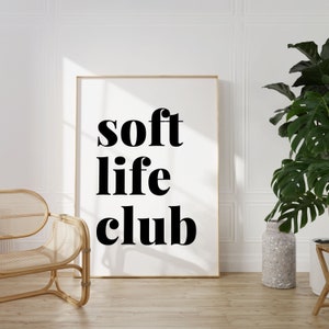 May include: A framed print with the words "soft life club" in bold black letters. The print is displayed in a room with a wooden floor, a woven chair, and a large potted plant. The frame is a light gold color.