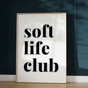 May include: A framed art print with the words "soft life club" in bold black letters on a white background. The frame is gold, and the print is displayed against a dark teal wall and light wood floor.