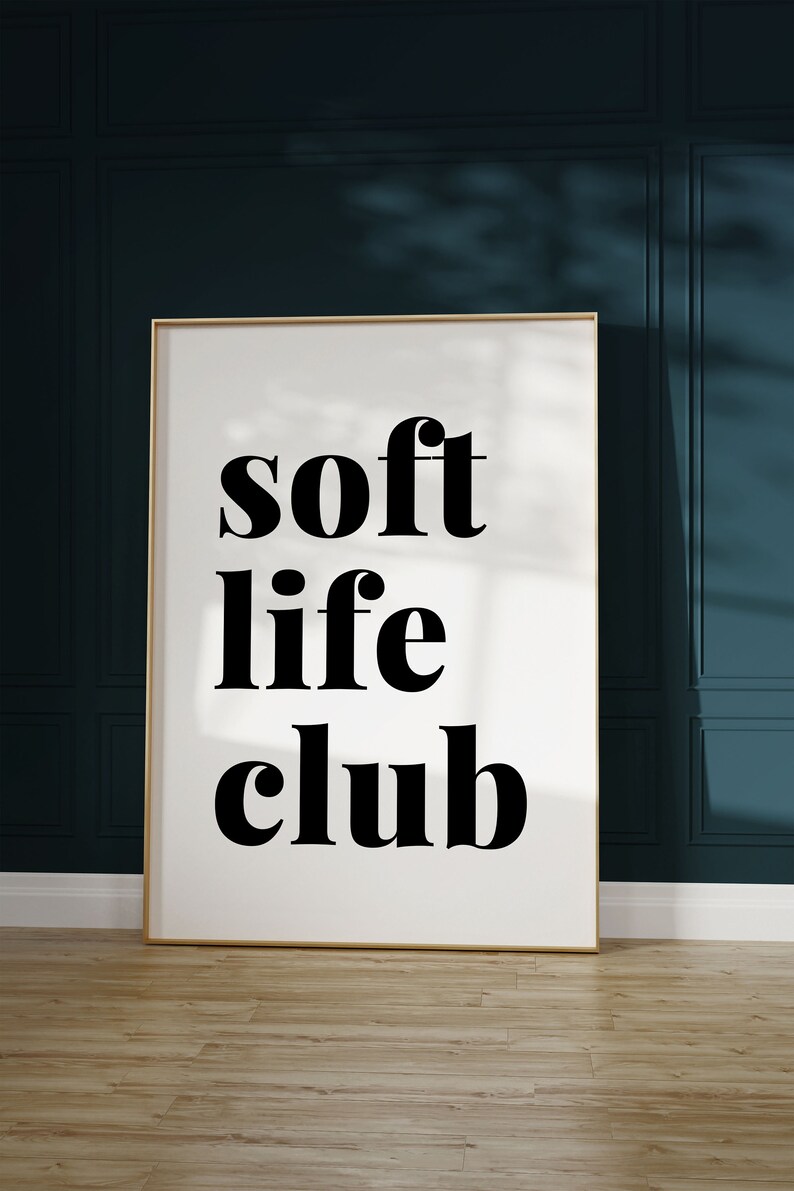 May include: A framed art print with the words "soft life club" in bold black letters on a white background. The frame is gold, and the print is displayed against a dark teal wall and light wood floor.