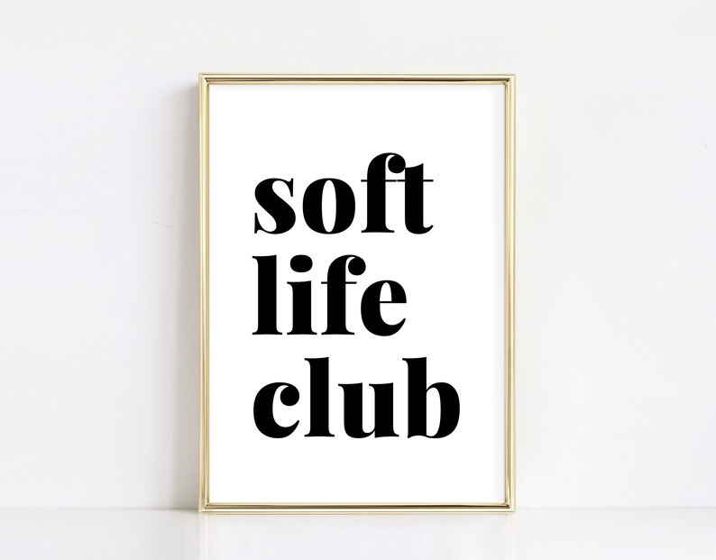 May include: A framed print with the words "soft life club" in bold black letters on a white background. The frame is gold. The print is displayed against a white wall.