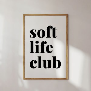 May include: A framed print with the words "soft life club" in bold black letters. The text is centered on a white background, encased in a thin, light brown wooden frame. The image is against a white wall.