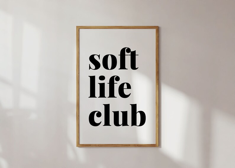 May include: A framed print with the words "soft life club" in bold black letters. The text is centered on a white background, encased in a thin, light brown wooden frame. The image is against a white wall.