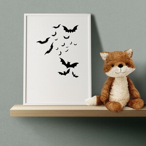 May include: A white picture frame with a black silhouette of bats on a white background. A brown plush fox toy is sitting on a wooden shelf in front of the frame.