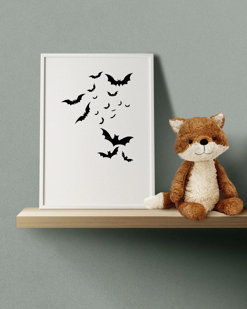 May include: A white picture frame with a black silhouette of bats on a white background. A brown plush fox toy is sitting on a wooden shelf in front of the frame.