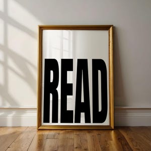 May include: A framed art print with the word "READ" in large, bold, black letters. The frame is gold-colored, and the print is leaning against a white wall on a wooden floor.