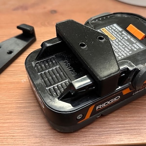 Op de afbeelding: Een zwarte en oranje Ridgid-accu met een zwarte plastic accuhouder. De accu heeft een label met de tekst "RIDGID 18V Lithium Ion Battery".