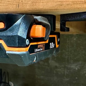 Op de afbeelding: Een zwarte en oranje 18V lithium-ion batterij is gemonteerd op een zwarte 3D-geprinte wandbevestiging. De batterij heeft een stroomindicator en een knop.