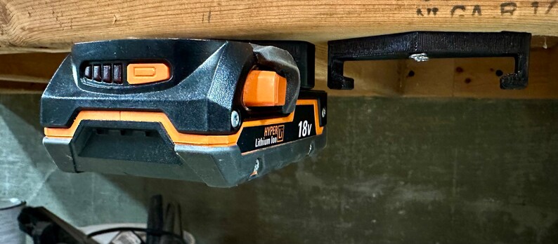 Op de afbeelding: Een zwarte en oranje 18V lithium-ion batterij is gemonteerd op een zwarte 3D-geprinte wandbevestiging. De batterij heeft een stroomindicator en een knop.