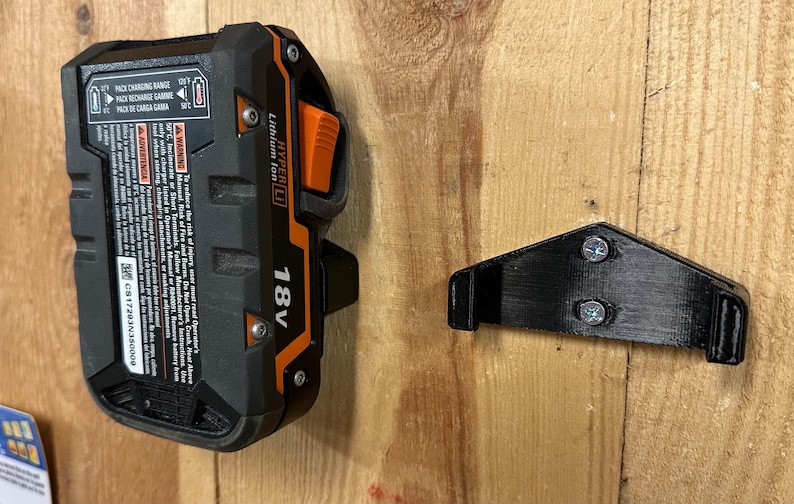 Op de afbeelding: Een zwarte en oranje 18V lithium-ion batterij met een zwarte plastic wandbevestiging. De batterij heeft een label met de tekst "HYPER LI" en "18V". De wandbevestiging heeft twee schroeven.
