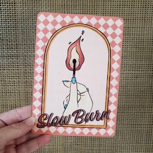May include: A rectangular card with a pink and white checkered border and a central illustration. The illustration depicts a hand holding a lit match with a flame. The text "Slow Burn" is written in a cursive font below the illustration.