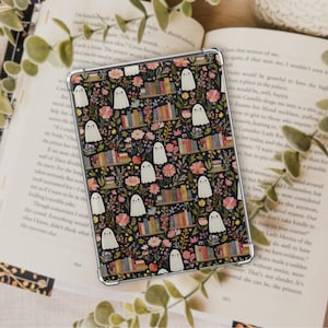 May include: A rectangular tablet case featuring a black background with a repeating pattern of small white ghosts, colorful floral arrangements, and miniature wooden bookshelves filled with various books arranged in rows across the surface.