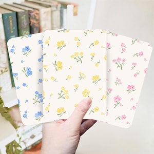May include: Three notebooks with floral patterns are held in a hand. Each notebook has a different colour scheme: blue, yellow, and pink flowers on a cream background. The notebooks are rectangular with rounded corners, and the floral design is repeated.