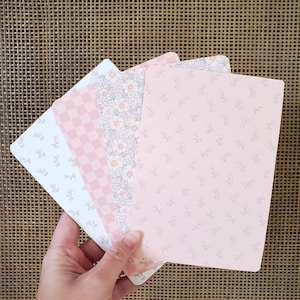 May include: A hand holding a stack of notebooks with floral patterns. The notebooks display designs such as small flowers, a checkered pattern, and a floral print, in shades of pink, white, and peach. The background has a woven texture.