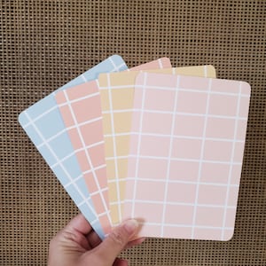 Op de afbeelding: Een hand houdt vier pastelkleurige kaarten vast. De kaarten zijn rechthoekig met afgeronde hoeken en een rasterpatroon. De kleuren zijn lichtblauw, perzik, geel en roze, met witte rasterlijnen. De achtergrond is een geweven textuur.