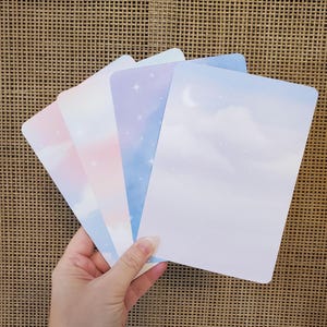 May include: A hand holding a fanned stack of rectangular cards with rounded corners. Each card features a different pastel sky-themed design, including clouds, stars, and a crescent moon. The colour palette includes pink, blue, and purple hues.
