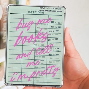 May include: A rectangular case with a light green library card design and the words "buy me books and tell me I'm pretty" in pink script. The case has a silver frame and is held in a hand.