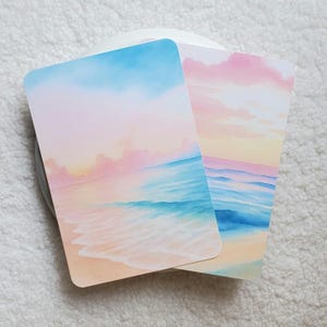 May include: Two watercolor art prints featuring beach scenes. One print has a blue sky and turquoise water, the other has a pink and yellow sky with blue water. Both prints have rounded corners and are on a white surface.