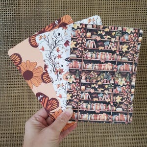 May include: Three decorative bookmarks with fall-themed designs. One bookmark features a floral and butterfly pattern in shades of orange and brown. Another has a tree branch and leaf design. The third bookmark displays a bookshelf.