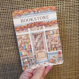 May include: A rectangular illustration of a bookstore with the word "BOOKSTORE" at the top. The storefront features a door and windows filled with books. Autumn leaves decorate the top. Stacks of books and a pumpkin are in front.