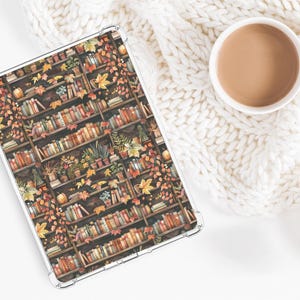 Puede incluir: Una funda transparente para tableta con un diseño de acuarela de una estantería llena de libros, plantas y hojas de otoño. Una taza blanca llena de una bebida marrón se encuentra sobre una manta blanca de punto.
